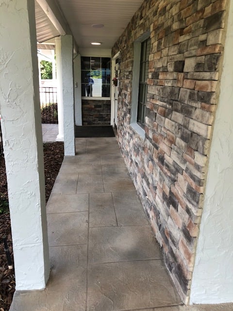 A stone-paved walkway with a brick wall and columns leading to a house entrance.