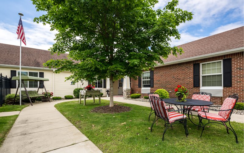 A cozy outdoor patio area with a table and chairs set under a tree, surrounded by a well-maintained lawn and brick house.
