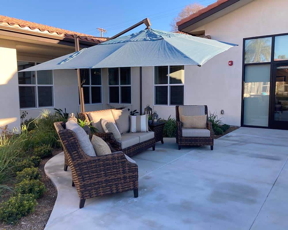 A cozy outdoor patio with rattan furniture and a large umbrella, set against a modern house backdrop.