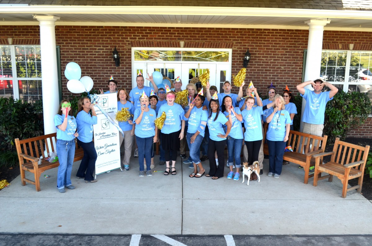 A group of people in blue shirts pose for a photo at an outdoor event with balloons and a banner.