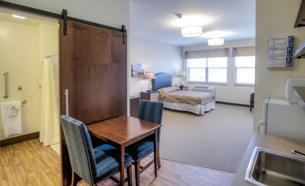 A spacious hotel room with a bed, table and chairs, featuring ample lighting and large windows.