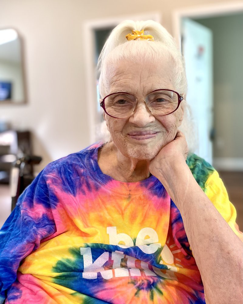 An elderly woman with glasses, wearing a colorful tie-dye shirt, smiles warmly indoors.