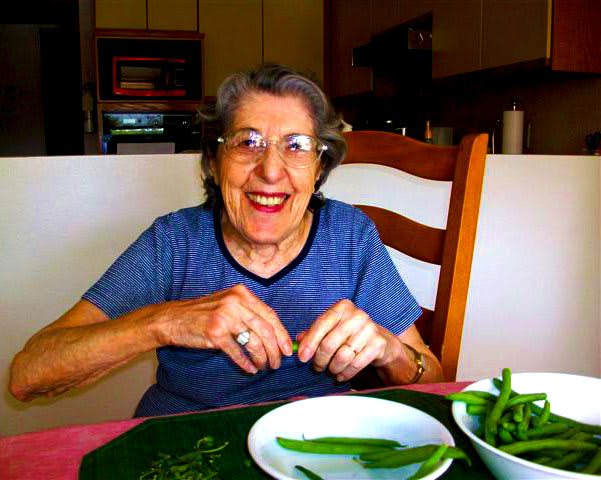 An elderly woman smiles while sitting at a dining table with vegetables.