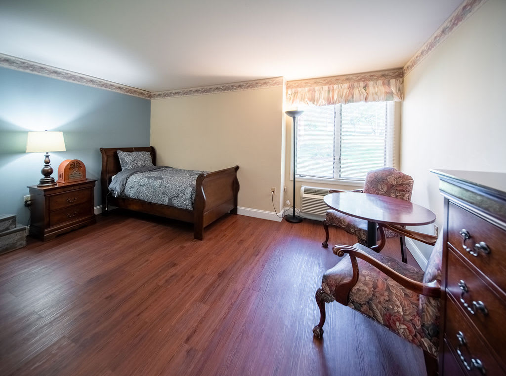 A spacious bedroom featuring a single wooden bed, a side table, and an upholstered chair next to a window.