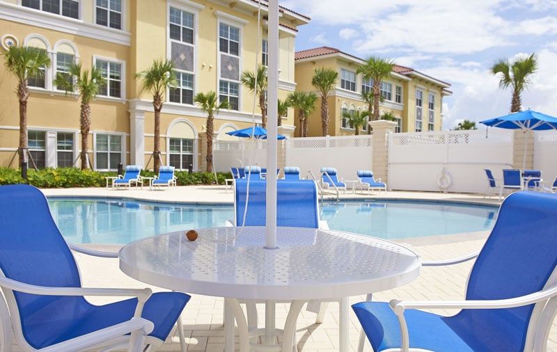 A sunny resort poolside area with blue chairs and tables, surrounded by palm trees.