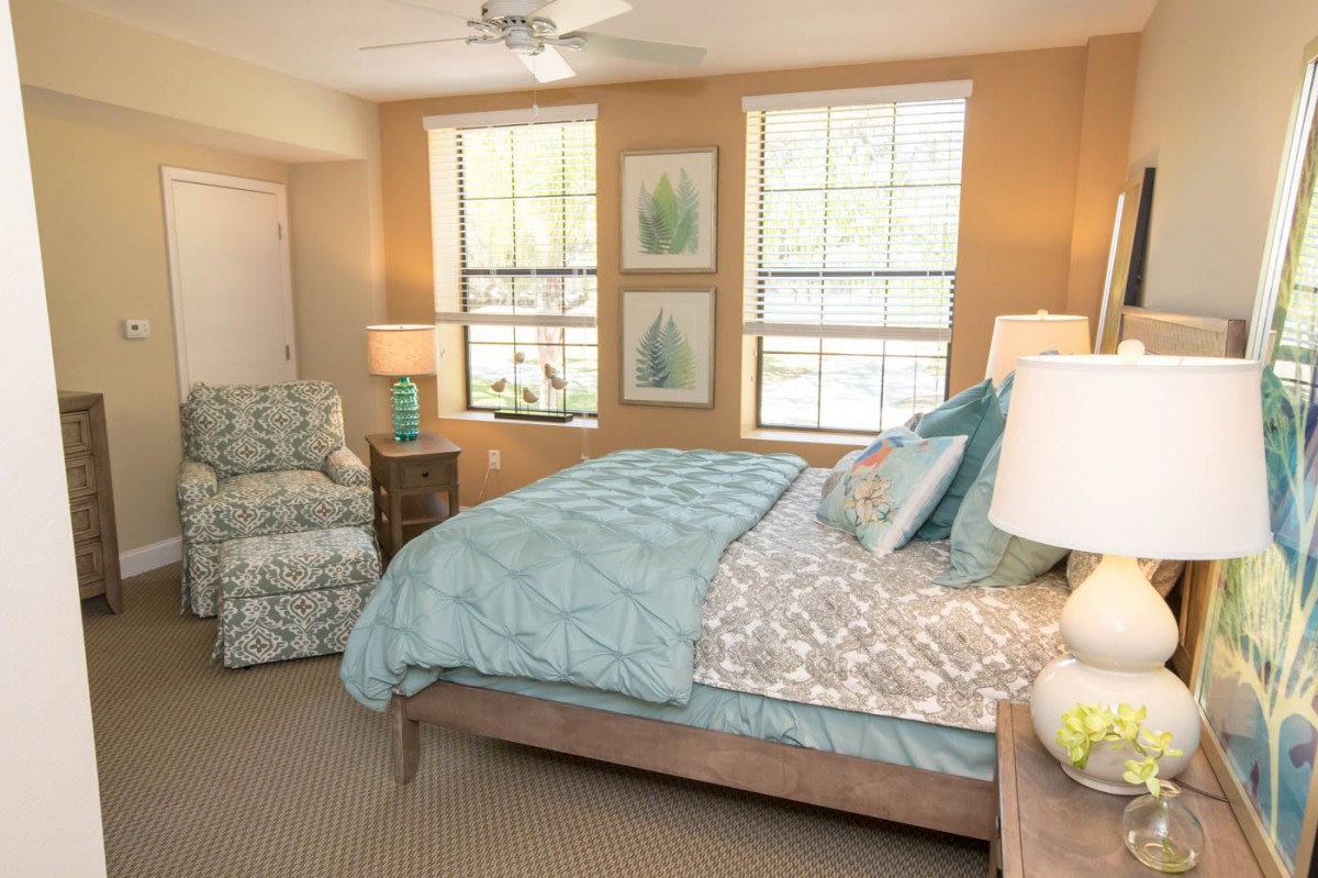 A cozy bedroom with a neatly made bed, an armchair, and large windows allowing natural light.
