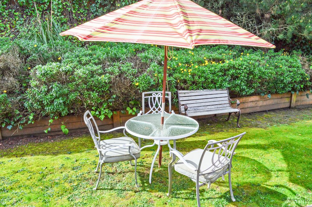 A garden scene featuring a patio set with a striped umbrella, surrounded by greenery.