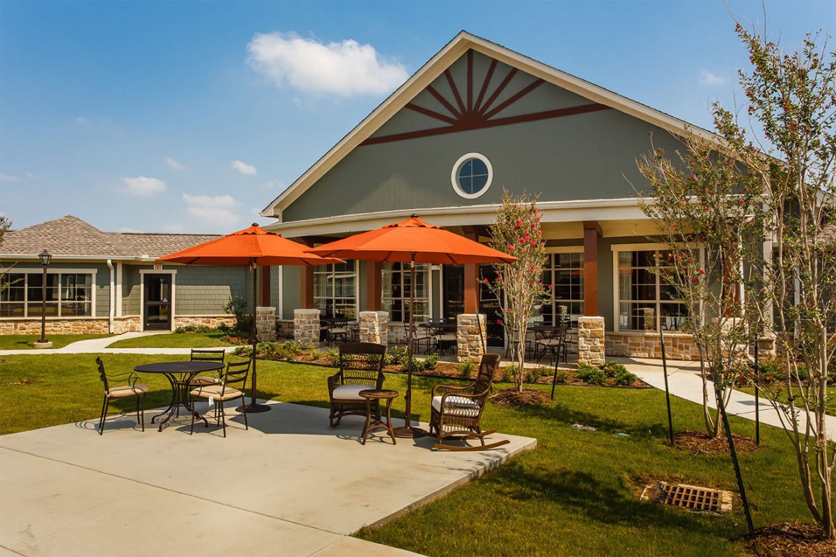 A residential building with a patio featuring tables, chairs, and orange umbrellas on a sunny day.