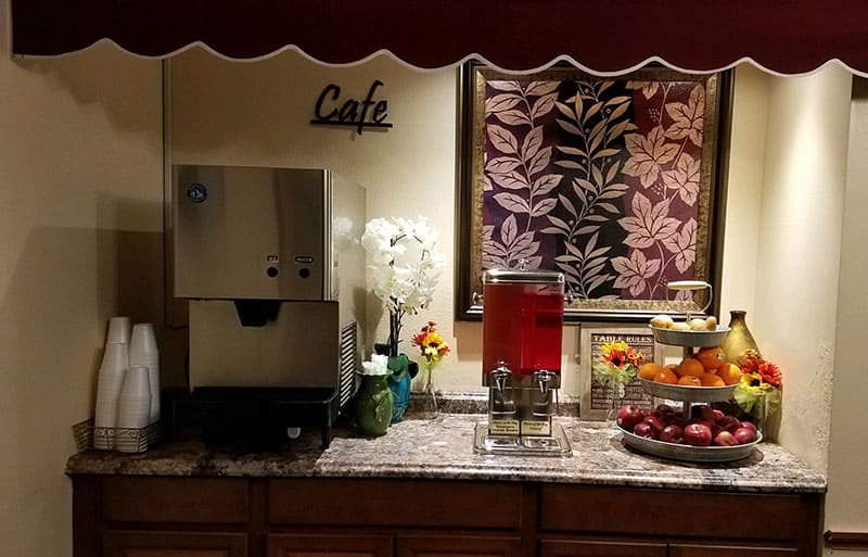 A cozy cafe corner featuring a beverage dispenser, coffee machine, fresh fruits, and decorative elements.