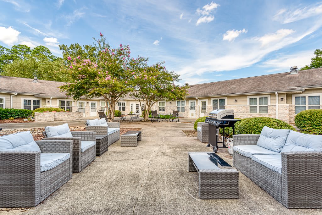 A spacious patio with comfortable seating and a grill, surrounded by greenery and a building under a blue sky.