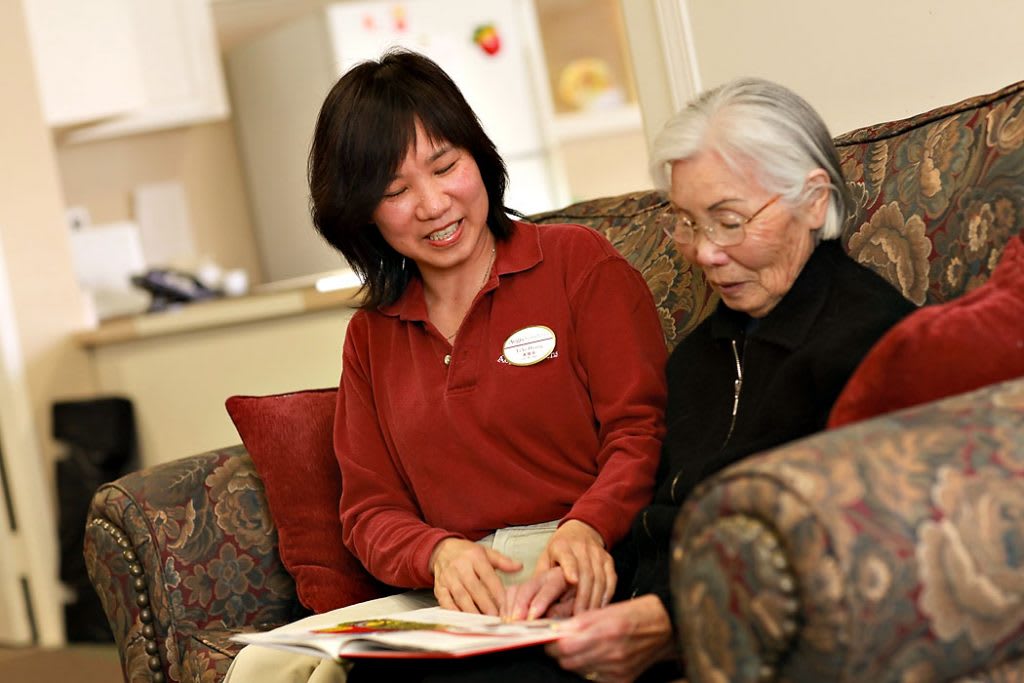 A caregiver reading with an elderly woman on a sofa.