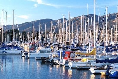 A bustling marina filled with sailboats against a backdrop of mountains and a clear blue sky.