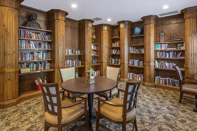 A cozy library room with wooden paneling, featuring bookshelves and a circular table surrounded by chairs.