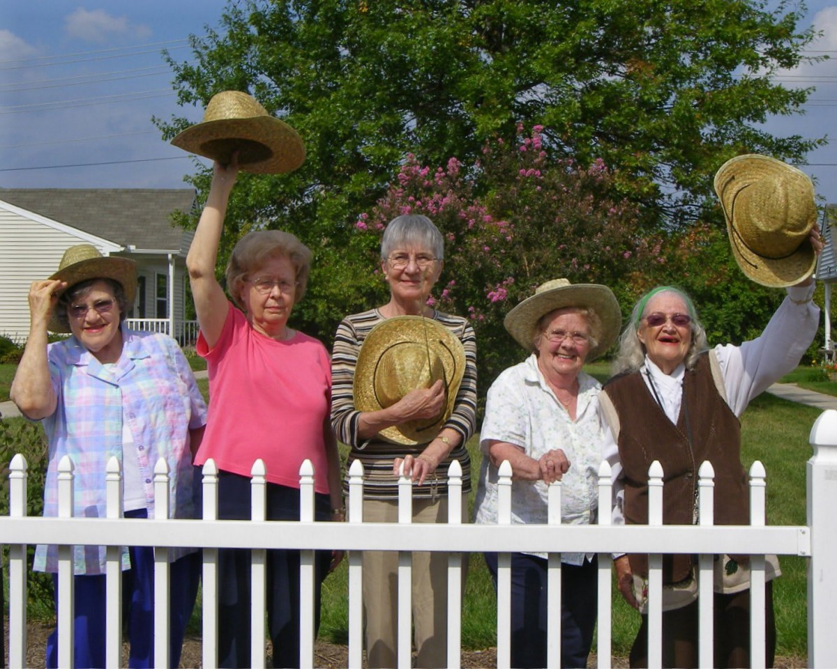 A group of women standing by a white fence, holding hats, enjoying a sunny day.