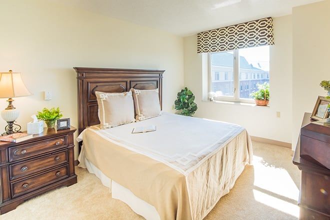 A bright and cozy bedroom with a neatly made bed, nightstand, window with patterned blinds, and potted plants.