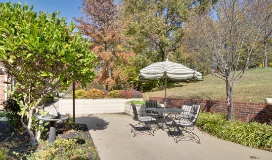 A serene outdoor patio area with a table, chairs, and umbrella surrounded by lush greenery and trees.