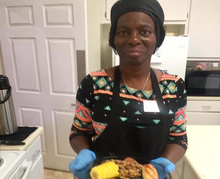 A woman in a kitchen wearing an apron and gloves, holding a plate of food.