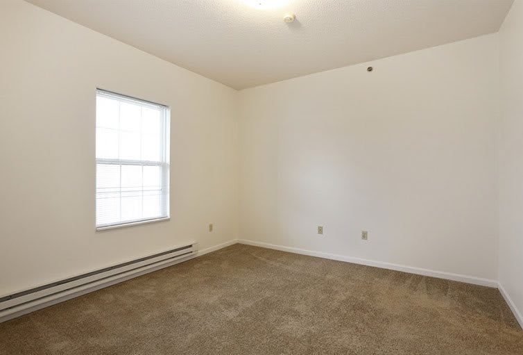 An empty room with white walls and a carpeted floor, featuring a window.