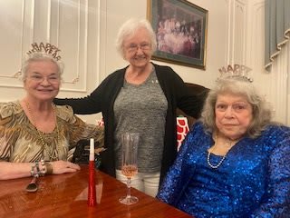 Three elderly women celebrating together, wearing tiaras and smiling at a party.
