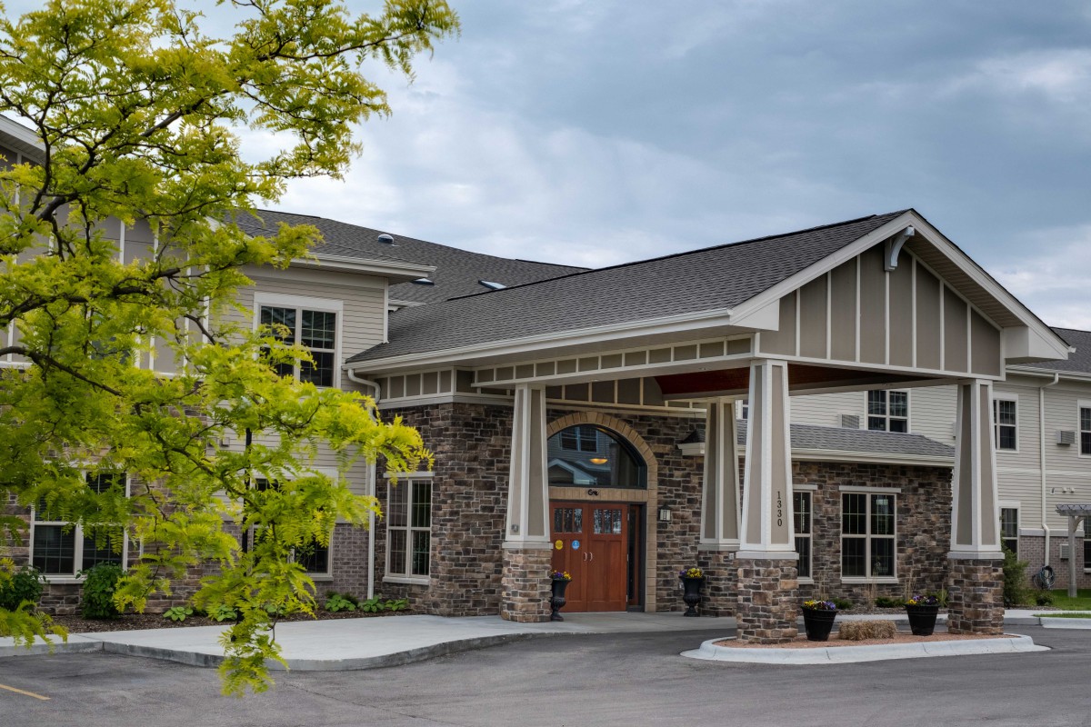 A modern building with a stone facade and a prominent entrance under a cloudy sky, surrounded by greenery.