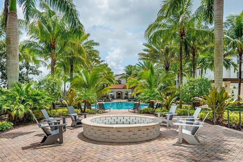 A tropical outdoor area featuring a fire pit surrounded by chairs with a pool in the background and lush palm trees.