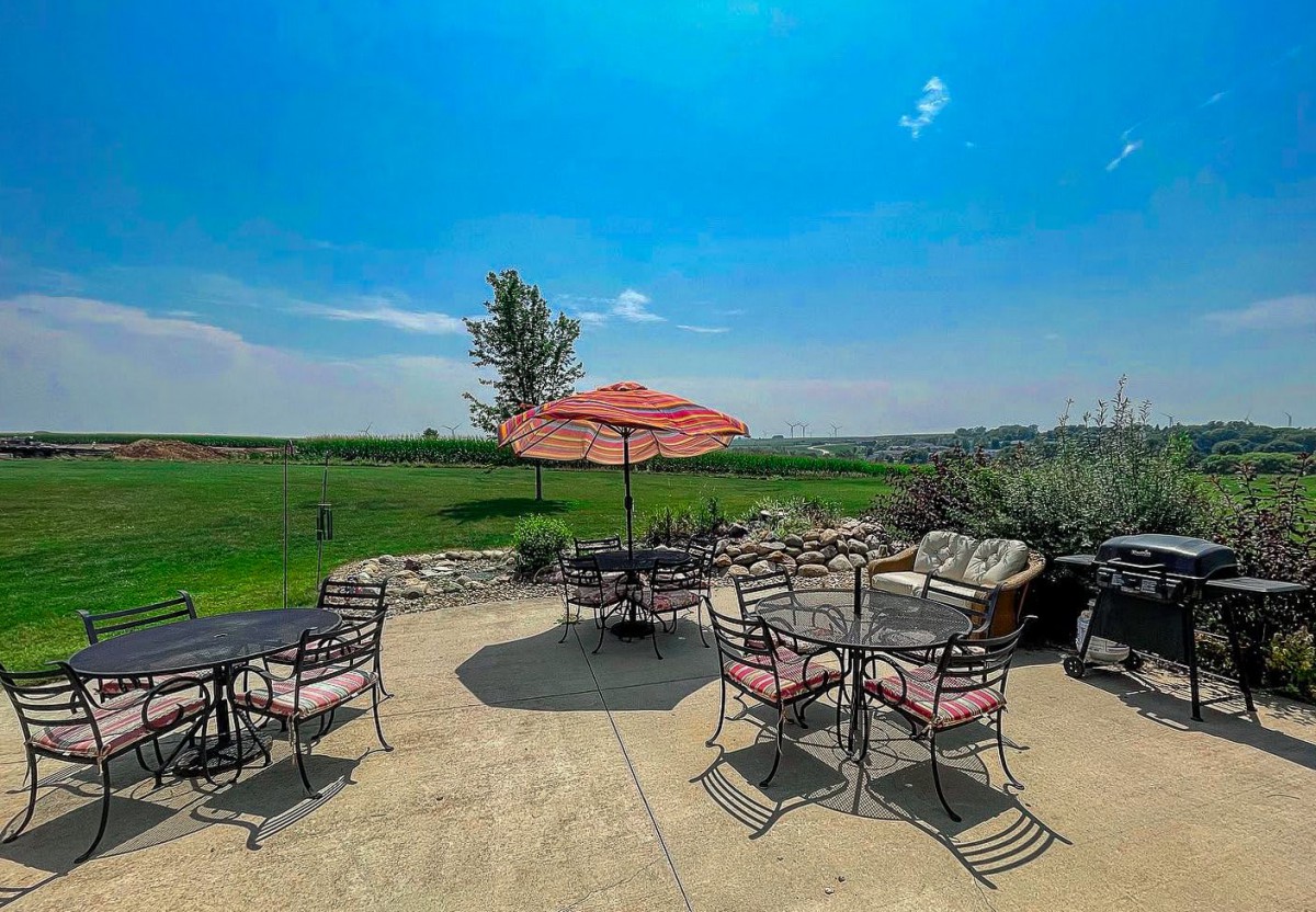 A sunny outdoor patio with tables, chairs, and an umbrella overlooking a green landscape.
