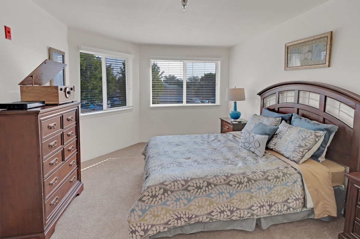 A cozy bedroom with a large bed, dresser, and two windows letting in natural light.