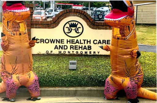 Two individuals in inflatable dinosaur costumes pose in front of Crowne Health Care and Rehab in Montgomery.