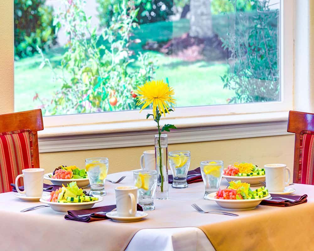 A beautifully set breakfast table with fresh fruit, cups, and a vase with a yellow flower by the window.