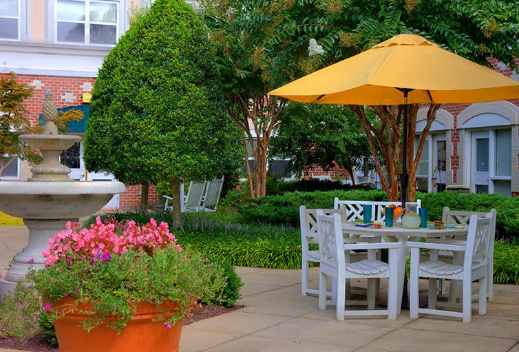 A cozy garden patio with white furniture and a yellow umbrella surrounded by lush greenery and vibrant flowers.
