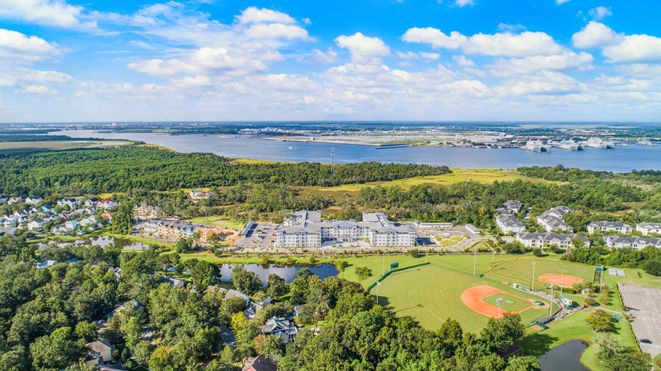 An aerial view of a landscape featuring buildings, a baseball field, and a river under a clear sky with clouds.