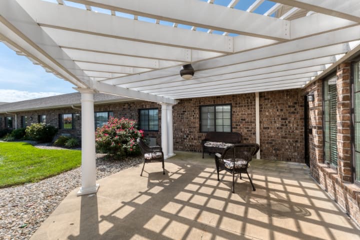 A sunny patio with a pergola casting shadows on the concrete, featuring outdoor seating against a brick wall.