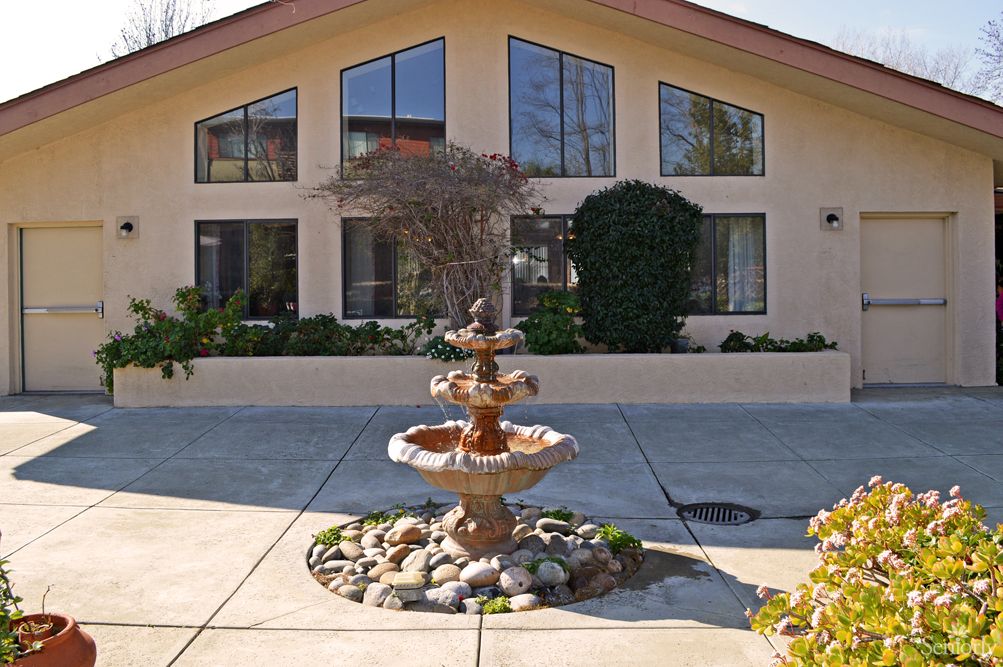 A three-tiered fountain stands in front of a building with large windows and a well-maintained garden.