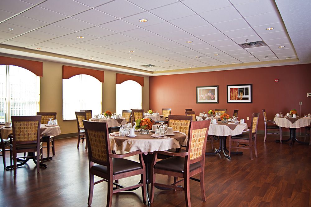 A cozy dining room with neatly set tables and chairs, featuring warm decor and natural light from large windows.