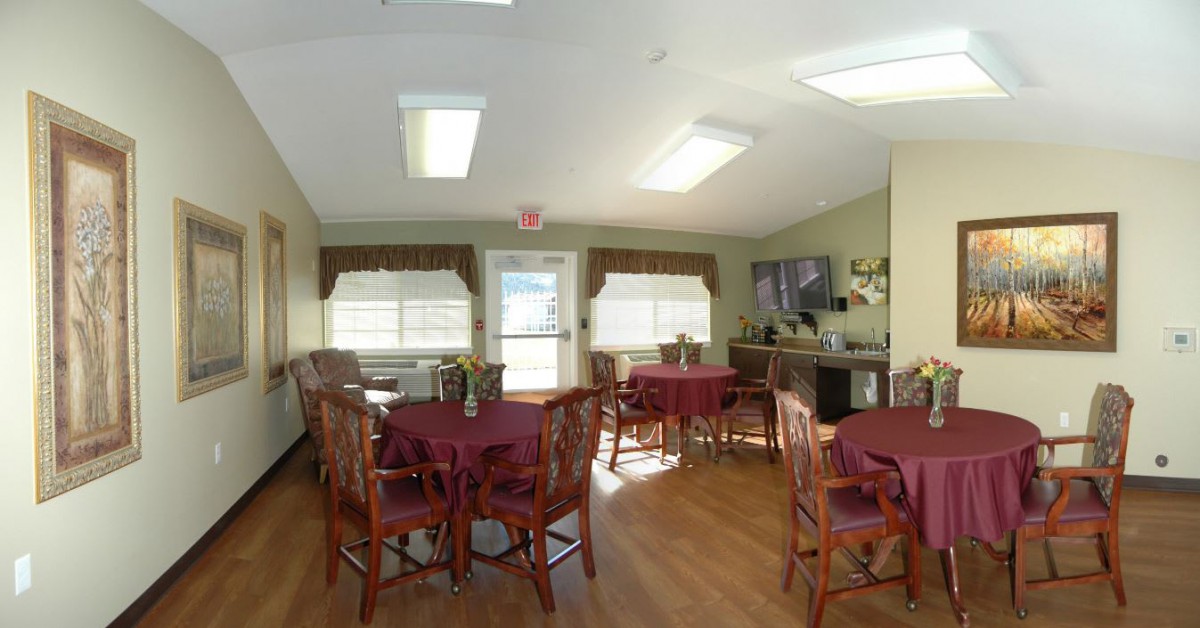 A well-lit dining area with multiple tables and chairs, featuring large windows and a wooden floor.