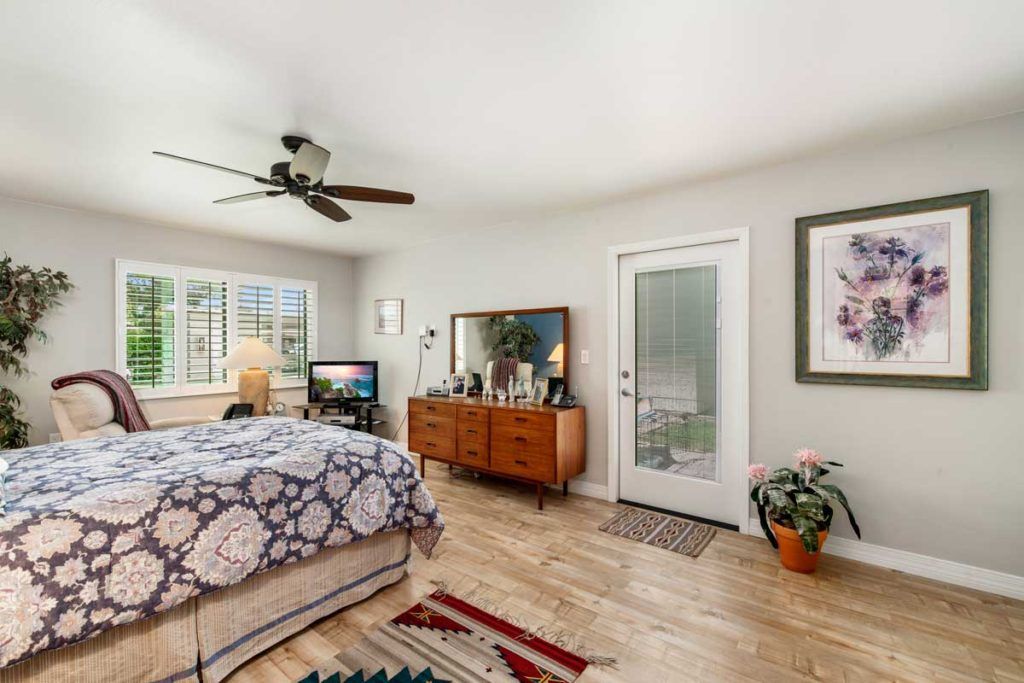 A cozy bedroom with a wooden floor, featuring a bed, dresser, ceiling fan, and abundant natural light.