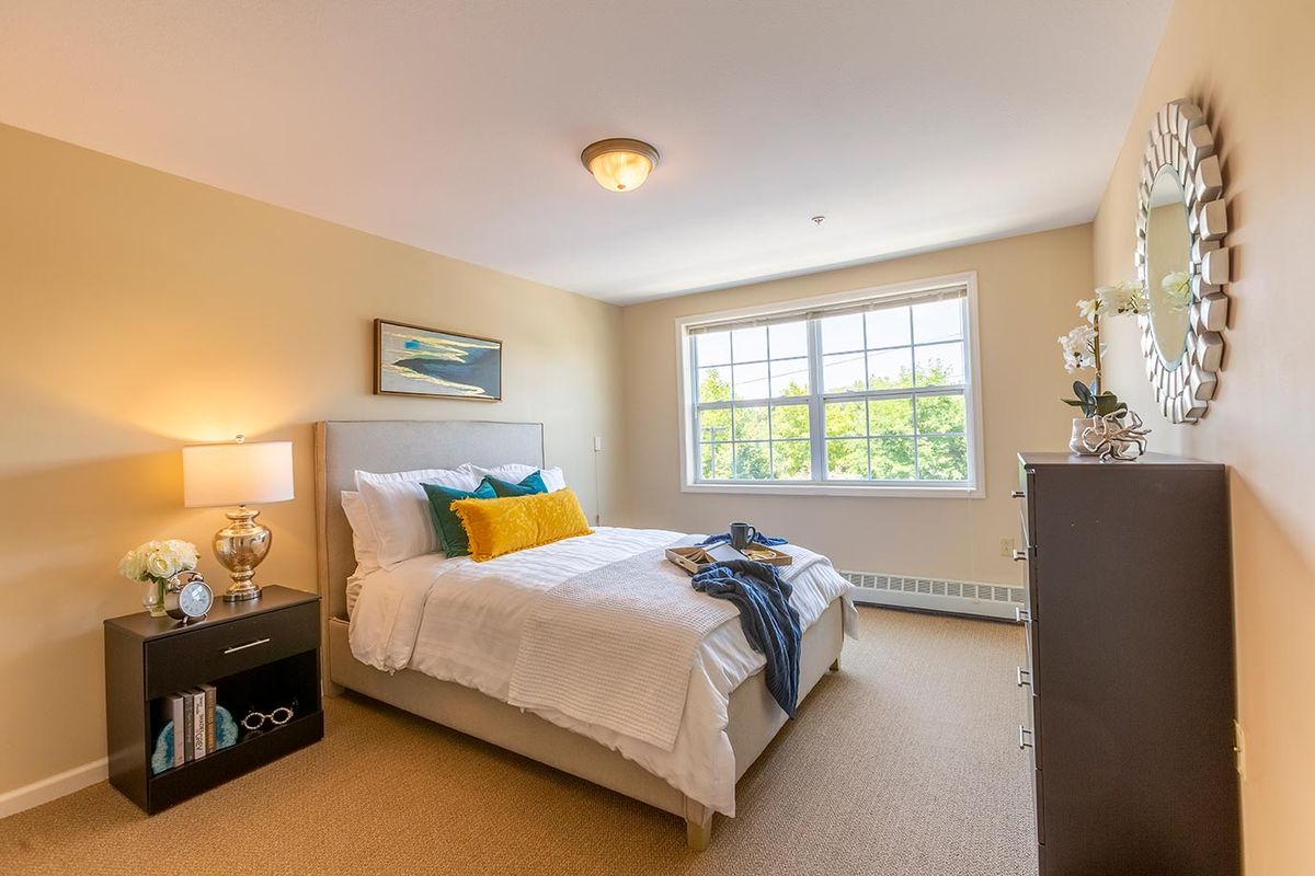 A cozy bedroom with a large window letting in natural light, featuring a neatly made bed with colorful pillows and simple decor.