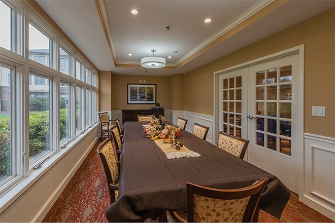 A well-decorated dining room with a long table and upholstered chairs, featuring large windows and ambient lighting.