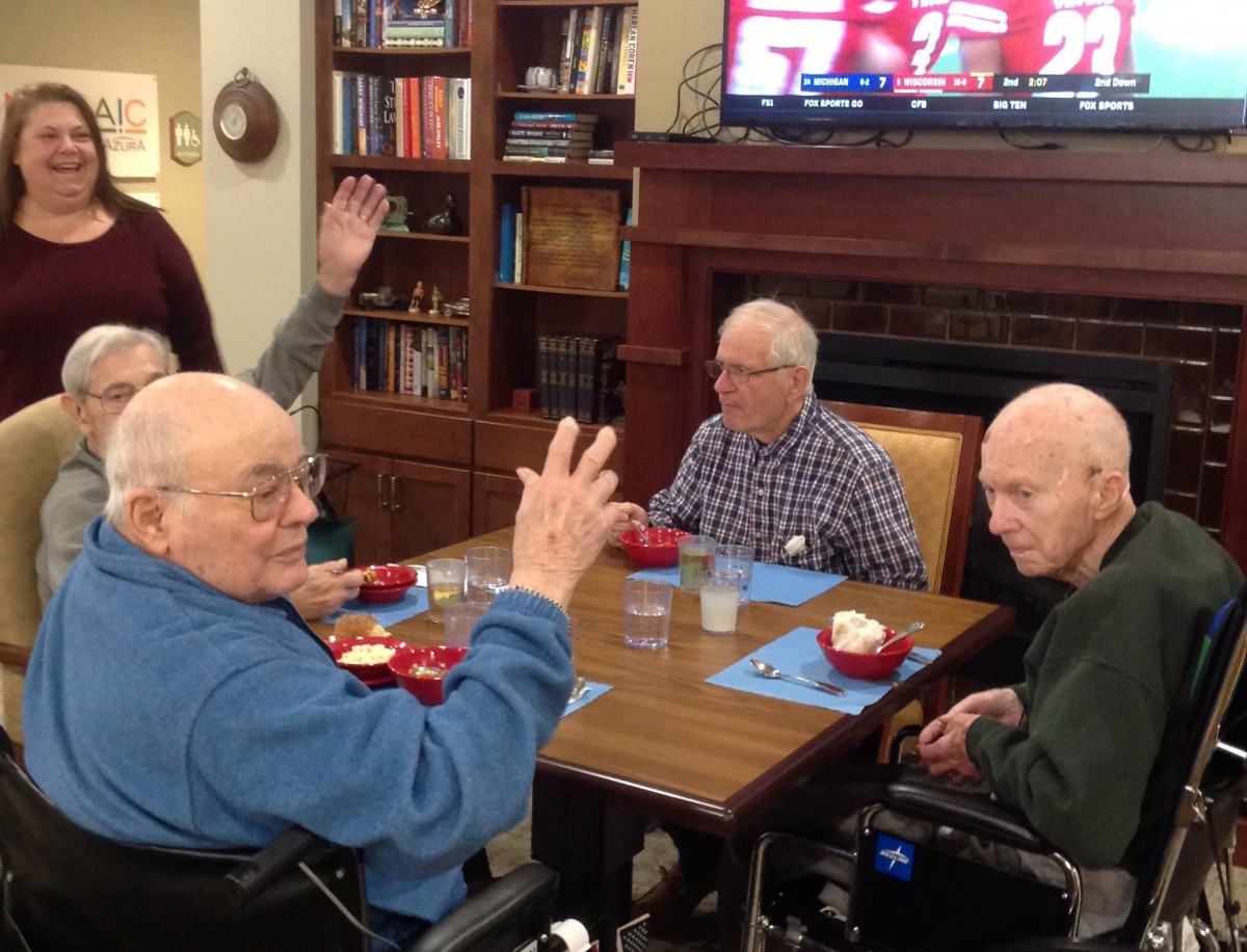 A group of seniors sitting around a table, engaging in conversation while watching TV.