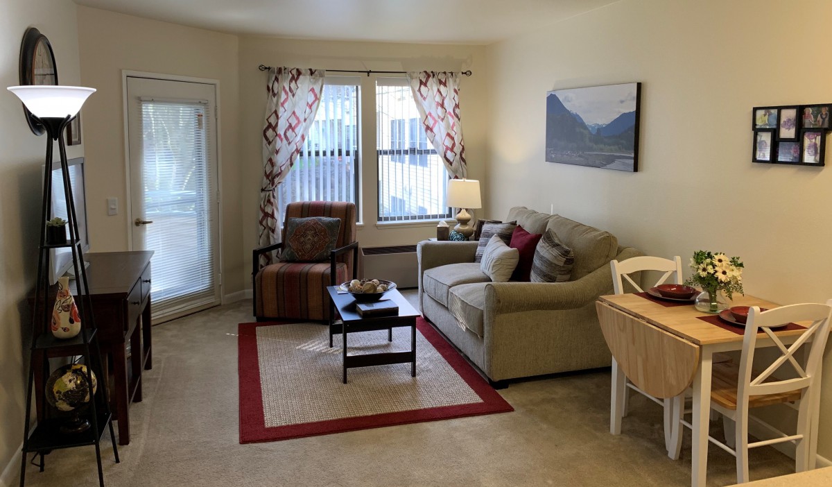 A cozy living room with a sofa, armchair, and coffee table arranged on a carpet, featuring artwork on the wall and a lamp near the window.