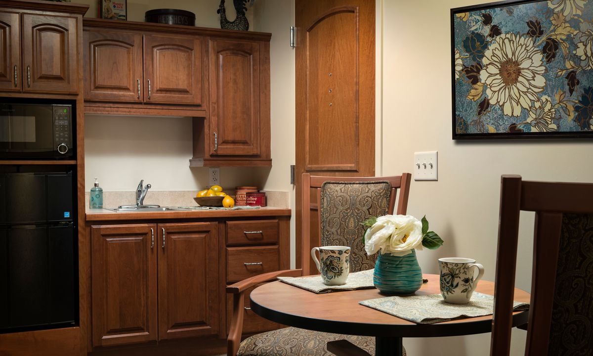 A cozy kitchen interior featuring wooden cabinets, a small dining table with flowers and mugs, and a bowl of fruit on the counter.