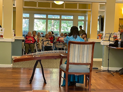 A performer plays music for an attentive audience of seniors in a welcoming indoor setting.