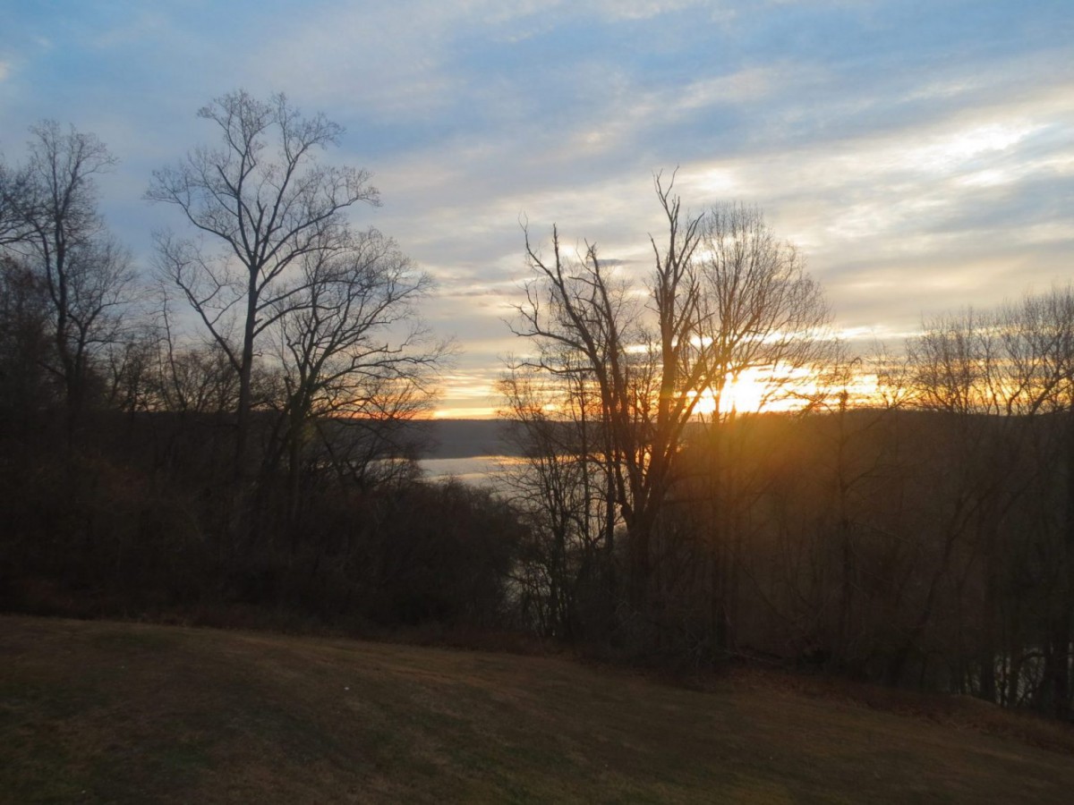 A serene sunset over a lake, framed by bare trees and a tranquil landscape.