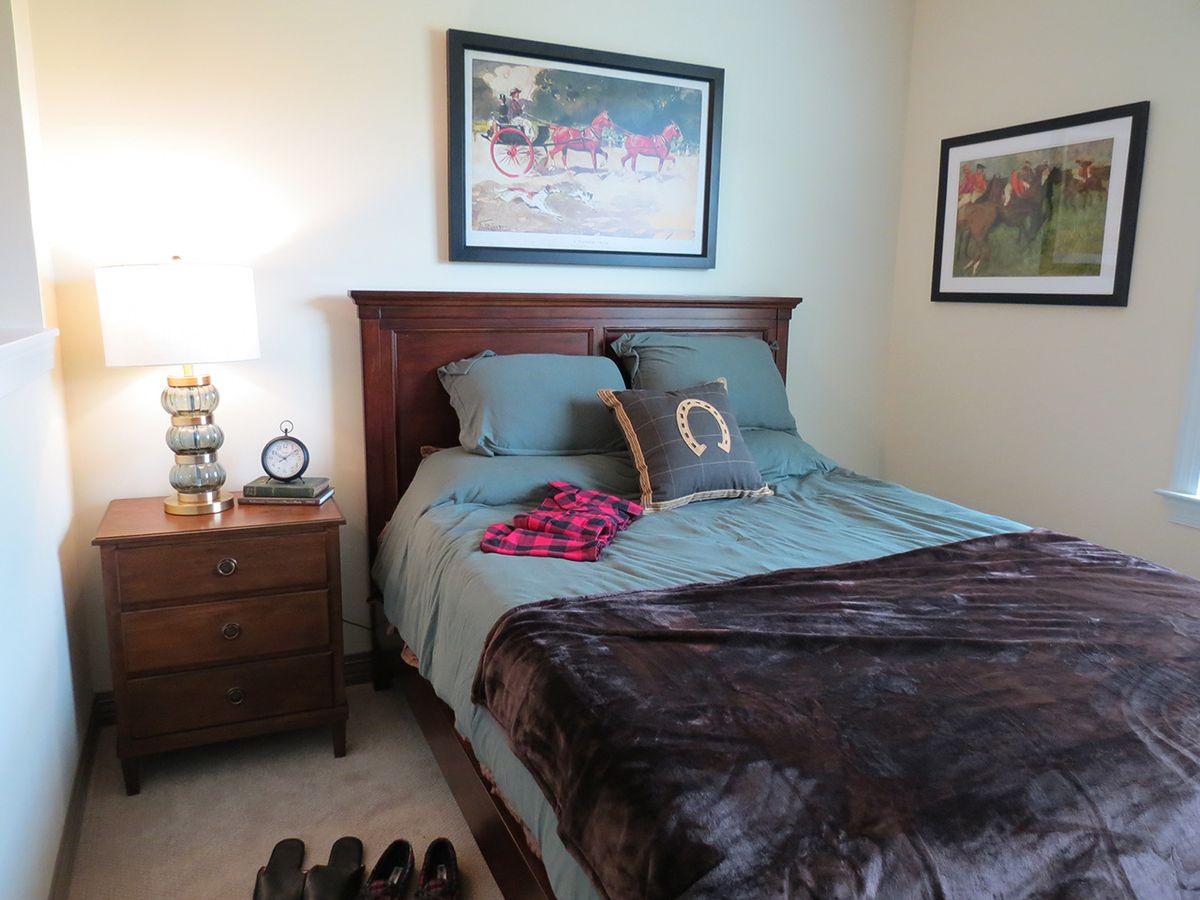 A cozy bedroom with a neatly made bed, featuring decorative pillows and a soft blanket. A nightstand with a lamp and alarm clock is on the side, and artwork adorns the walls.