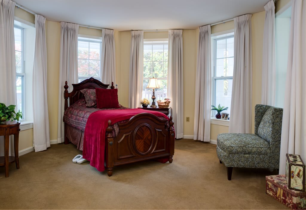 A cozy bedroom with a traditional wooden bed and red bedding, featuring bay windows with curtains.