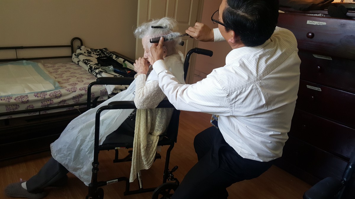 A caregiver is giving an elderly person a haircut while they sit in a wheelchair indoors.