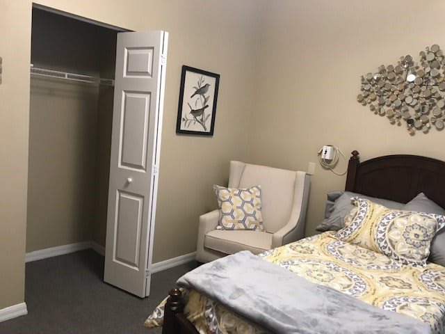 A cozy bedroom with a neatly made bed, upholstered chair, open closet door, and decorative wall art.
