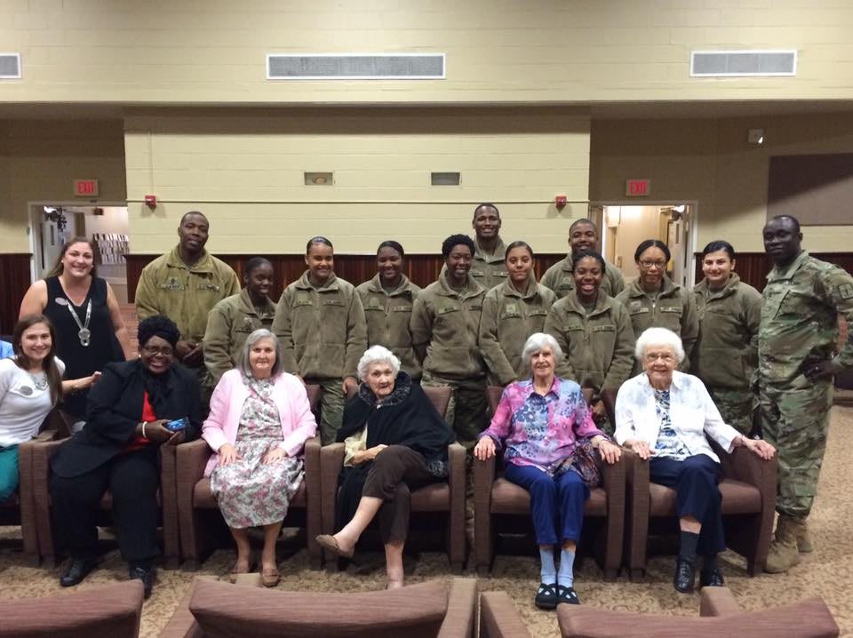 Military personnel pose with elderly women in a group photo indoors.