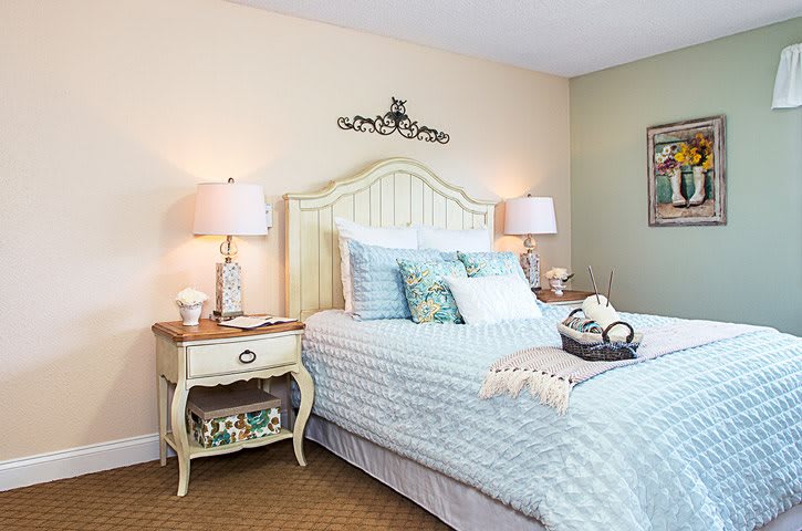 A cozy bedroom with pastel tones, featuring a bed with decorative pillows, nightstands with lamps, and stylish decor.
