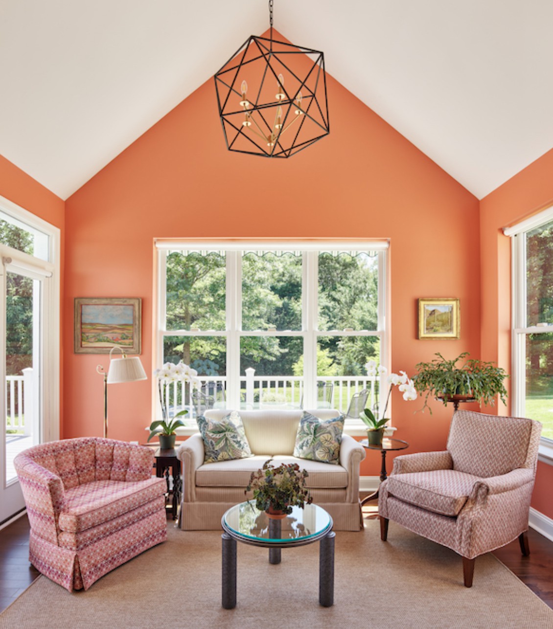 A cozy living room with orange walls, featuring a sofa and armchairs around a coffee table, with large windows overlooking a garden.